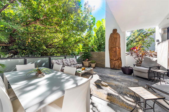 Outdoor patio area with seating, dining table, and large wooden sculpture surrounded by lush vegetation.