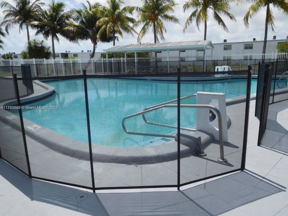 Outdoor swimming pool surrounded by a safety fence with palm trees in the background.