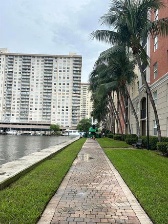View of a waterfront apartment building with a paved walkway and palm trees along the side.