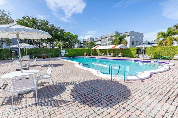 Outdoor swimming pool with deck chairs and umbrellas, surrounded by hedges and buildings.