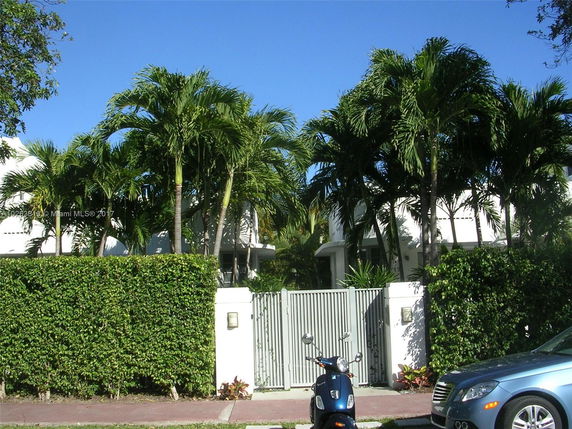 Front view of a house partially obscured by palm trees and a high hedge.