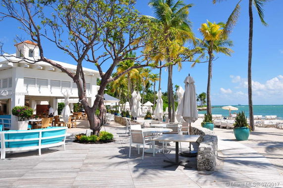 Outdoor seating area next to a beach with a view of palm trees and a white building.