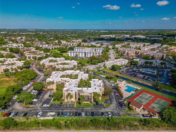 Aerial view showing multiple residential buildings with flat roofs. Nearby are parking lots, a swimming pool, and tennis courts, surrounded by greenery and roads.