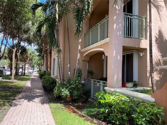 Front view of a multi-story building with balconies and a walkway lined with plants and trees.