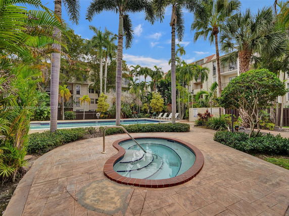 Outdoor area with swimming pool and hot tub surrounded by palm trees and apartment buildings.