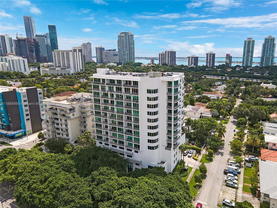 Aerial view of a tall white residential building in an urban area, surrounded by trees and other high-rise buildings.