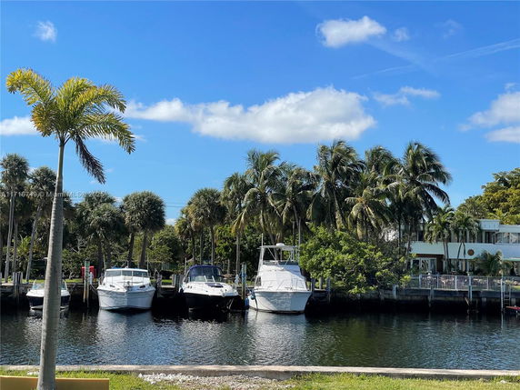 View of boats docked along a waterfront with palm trees in the background and a residential building nearby.