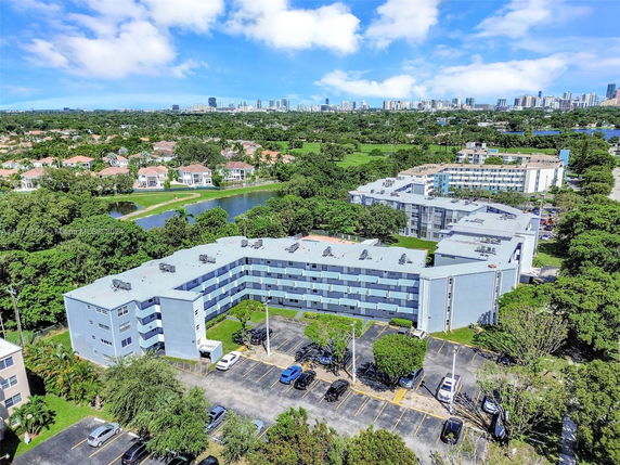 Aerial view of a four-story apartment complex with parking and greenery, surrounded by residential houses and distant city skyline.