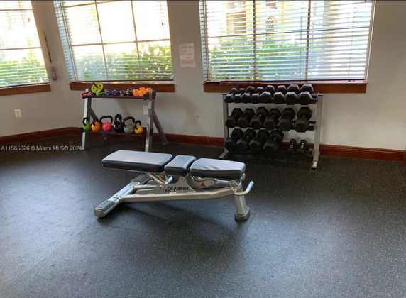 Interior view of a gym area with a bench press, dumbbells, and kettlebells near windows.