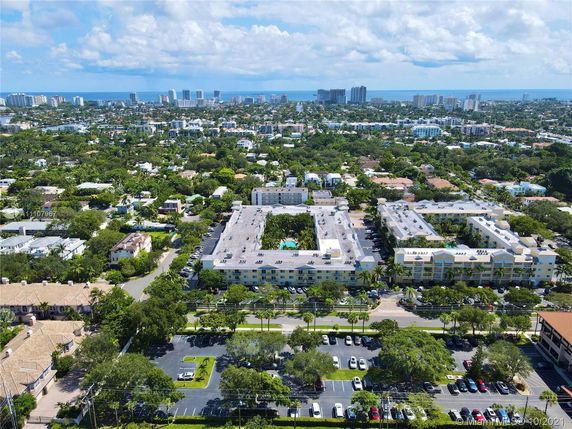 Aerial panoramic view of a residential area with buildings and green spaces, leading to a distant city skyline near the ocean.