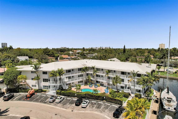 Elevated view of a multi-story building with a swimming pool at its center, surrounded by palm trees and parking area.
