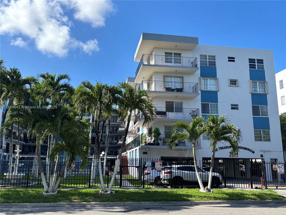 Front view of a multi-story condominium building with balconies and palm trees in front.