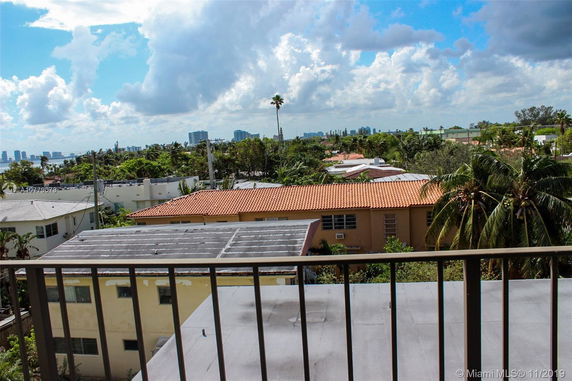 View from a balcony showing rooftops with orange tiles, surrounding green areas, and a city skyline in the distance.