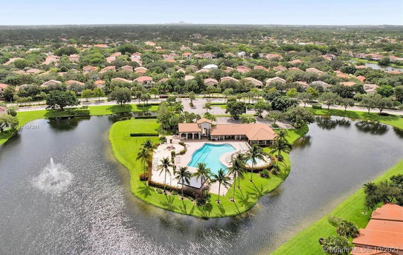 Panoramic view of a residential community with a clubhouse and swimming pool, surrounded by greenery and a water feature.