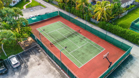 Aerial view of a tennis court with surrounding fence and nearby parking area.