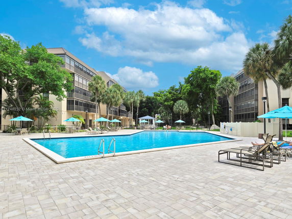 Outdoor swimming pool area with sun loungers and umbrellas near a multi-story building.