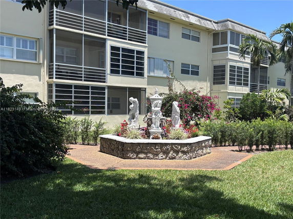 Front view of a multi-story residential building with enclosed balconies and a garden area with statues.