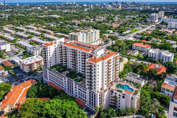 Aerial view of a multi-story residential building with a swimming pool on a higher floor, surrounded by lush greenery and other buildings in an urban area.
