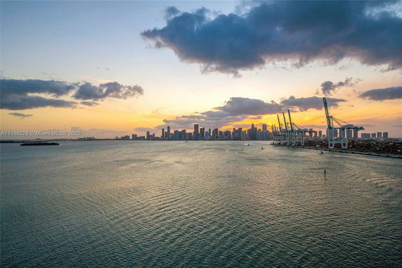 Skyline view of a city at sunset with water and port cranes.