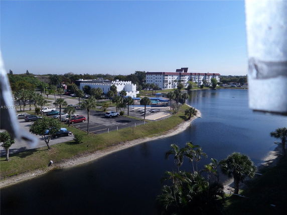 View of several buildings by a lake with a parking area and palm trees.