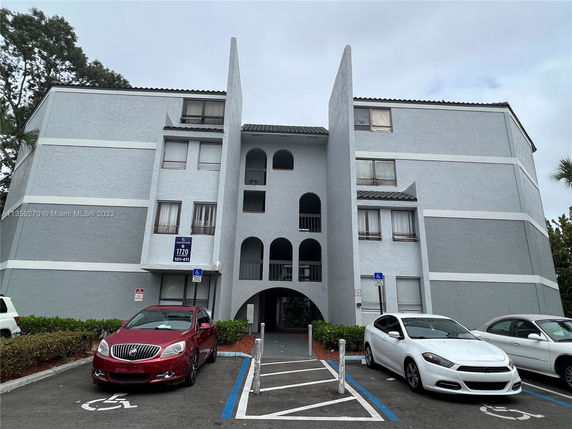 Front view of a multi-story apartment building with arched balconies and parking spaces in front.