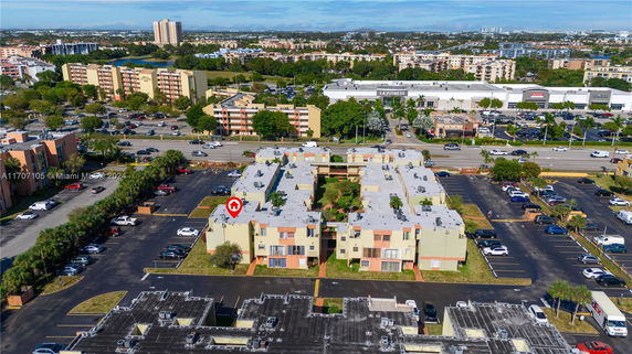 Aerial view of a large residential complex with multiple buildings and parking areas, surrounded by a busy road and various commercial establishments in the background.