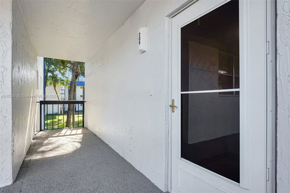Exterior view of a building corridor with a glass door and railing, leading outside with trees visible.
