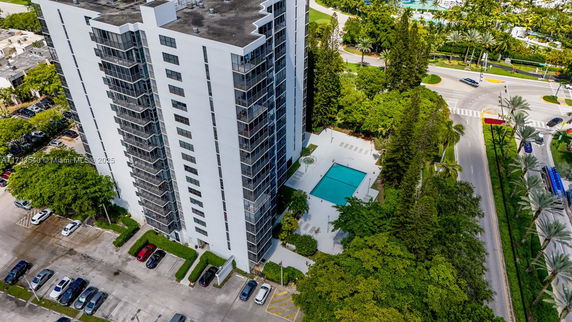 Aerial view of a high-rise building with a visible swimming pool and parking area surrounded by greenery and roads.