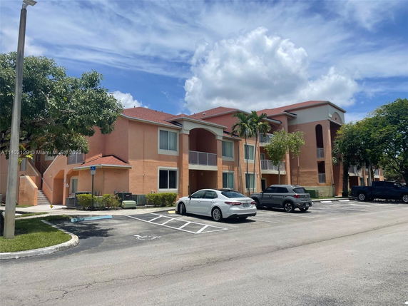 Front view of a multi-story apartment building with balconies and a parking area in front.
