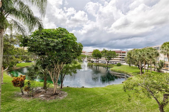 Scenic view of a pond with fountain, surrounded by trees and buildings in the background.