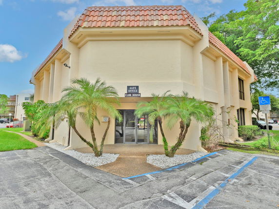 Front view of a two-story clubhouse with a tiled roof and palm trees at the entrance.
