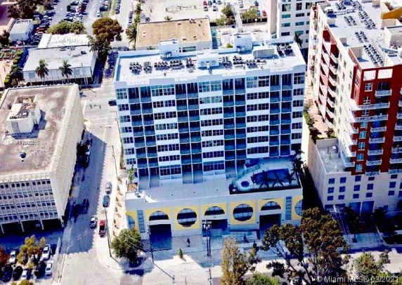 Aerial view of a multi-story building with a flat roof and balconies. Surrounding buildings and streets are visible.