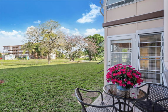 View from a patio with outdoor seating and a potted plant, overlooking a grassy area with trees and a building in the background.