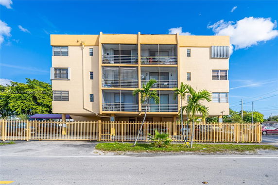 Front view of a four-story apartment building with balconies and a fenced area.