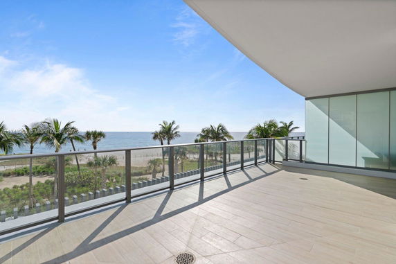 Wide balcony with glass railing offering a view of the ocean and palm trees.