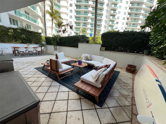 Outdoor patio area of an apartment with seating and dining furniture, surrounded by greenery and adjacent to a building with balconies.