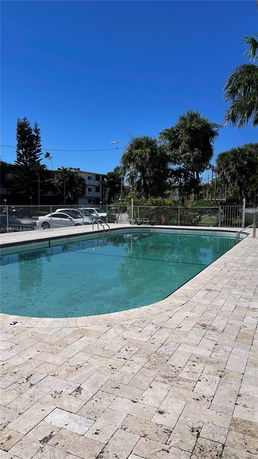Outdoor swimming pool surrounded by a paved area with trees and buildings in the background.
