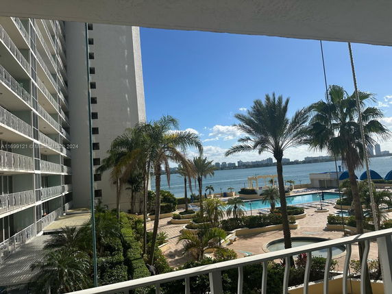 View from a building showing a swimming pool area with palm trees, overlooking a body of water and distant city skyline.