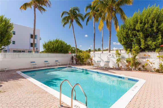 Outdoor pool area with lounge chairs and palm trees. A multi-story building is visible in the background.