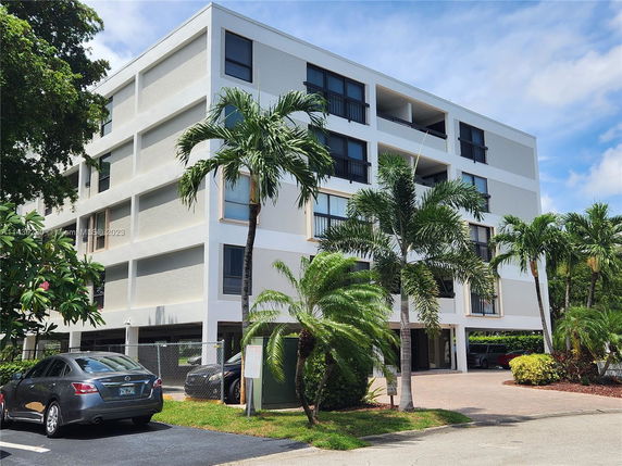 Front view of a four-story apartment building with balconies and surrounding palm trees.
