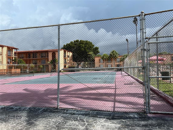 View of an outdoor tennis court enclosed by a chain-link fence with residential buildings in the background.