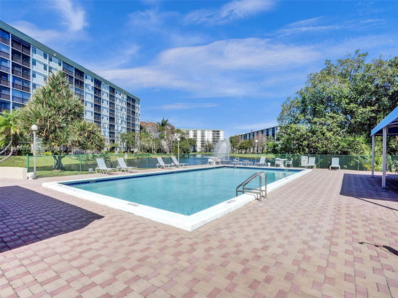Outdoor swimming pool area with surrounding lounge chairs and apartment buildings in the background.