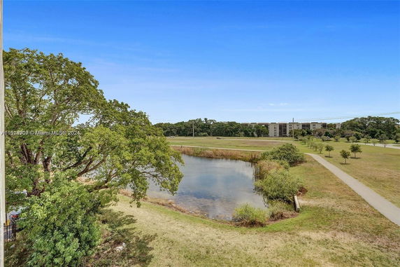 View of a grassy area with a pond and trees, and buildings in the distance.