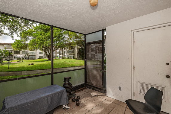 Covered outdoor area with tiled floor, overlooking grassy courtyard and nearby building.