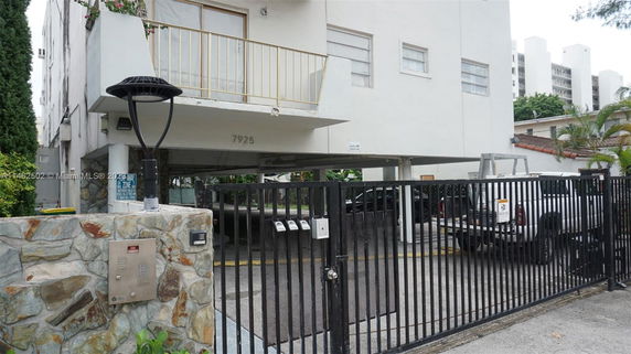 View of a building with a gated entrance to a parking area beneath a raised structure.