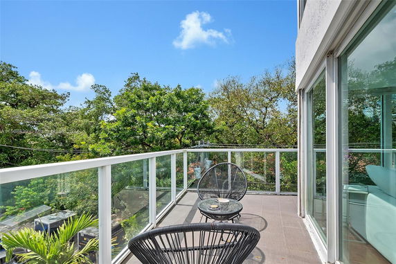 Outdoor balcony with glass railings and chairs, overlooking green trees.