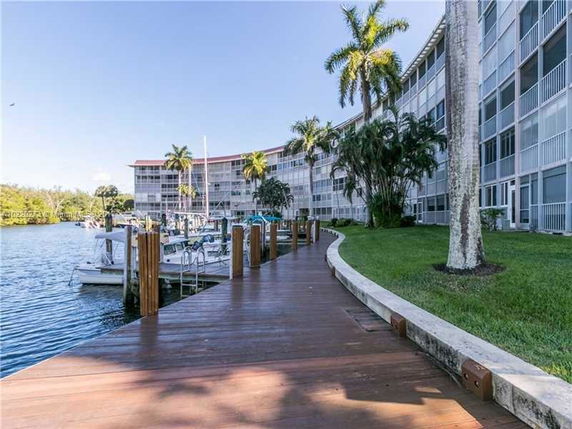 View of a waterfront residential building with multiple floors and balconies, surrounded by palm trees. A wooden dock is seen in the foreground next to the water with boats docked.