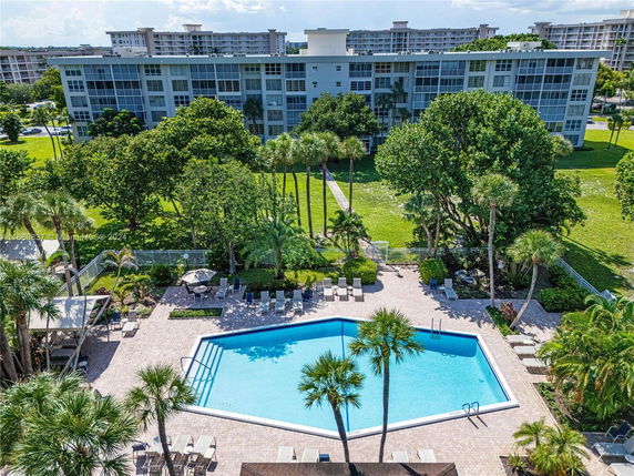 A large residential building with a swimming pool area in the foreground surrounded by trees and seating.