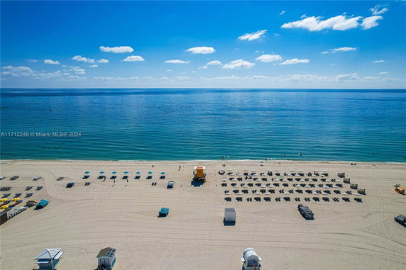 View of a sandy beach with rows of beach chairs and umbrellas overlooking a calm blue ocean.