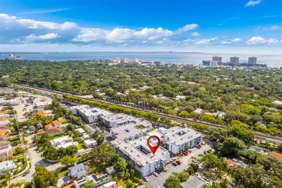Aerial view of a residential area with multiple buildings, surrounded by greenery, with a water body and cityscape in the background.